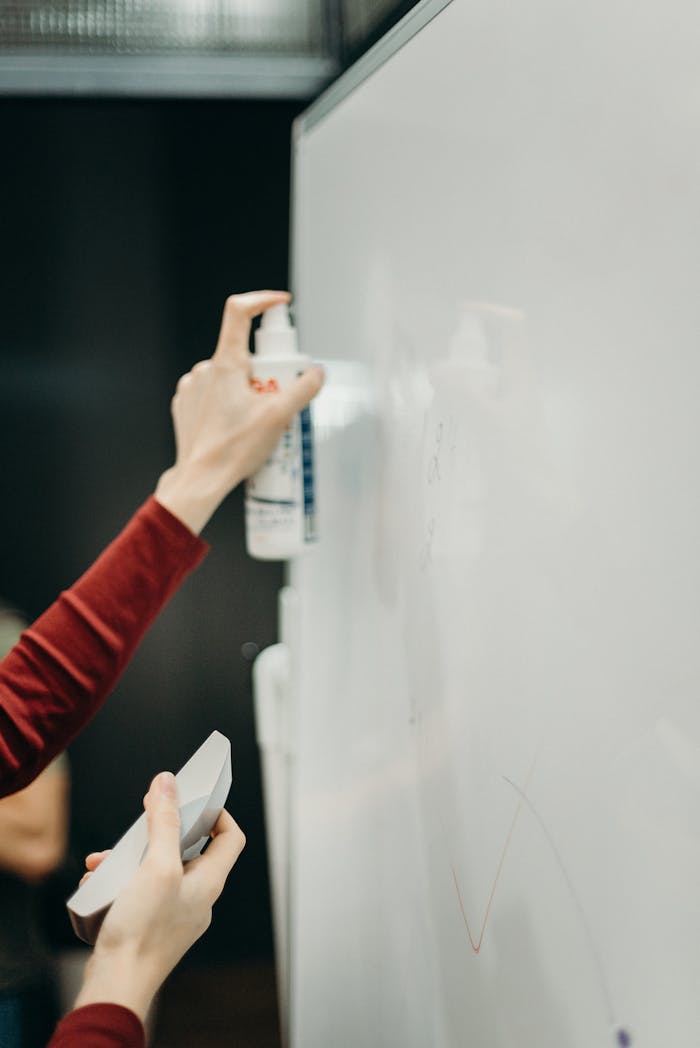 Close-up of hands cleaning a whiteboard with spray bottle in an office.