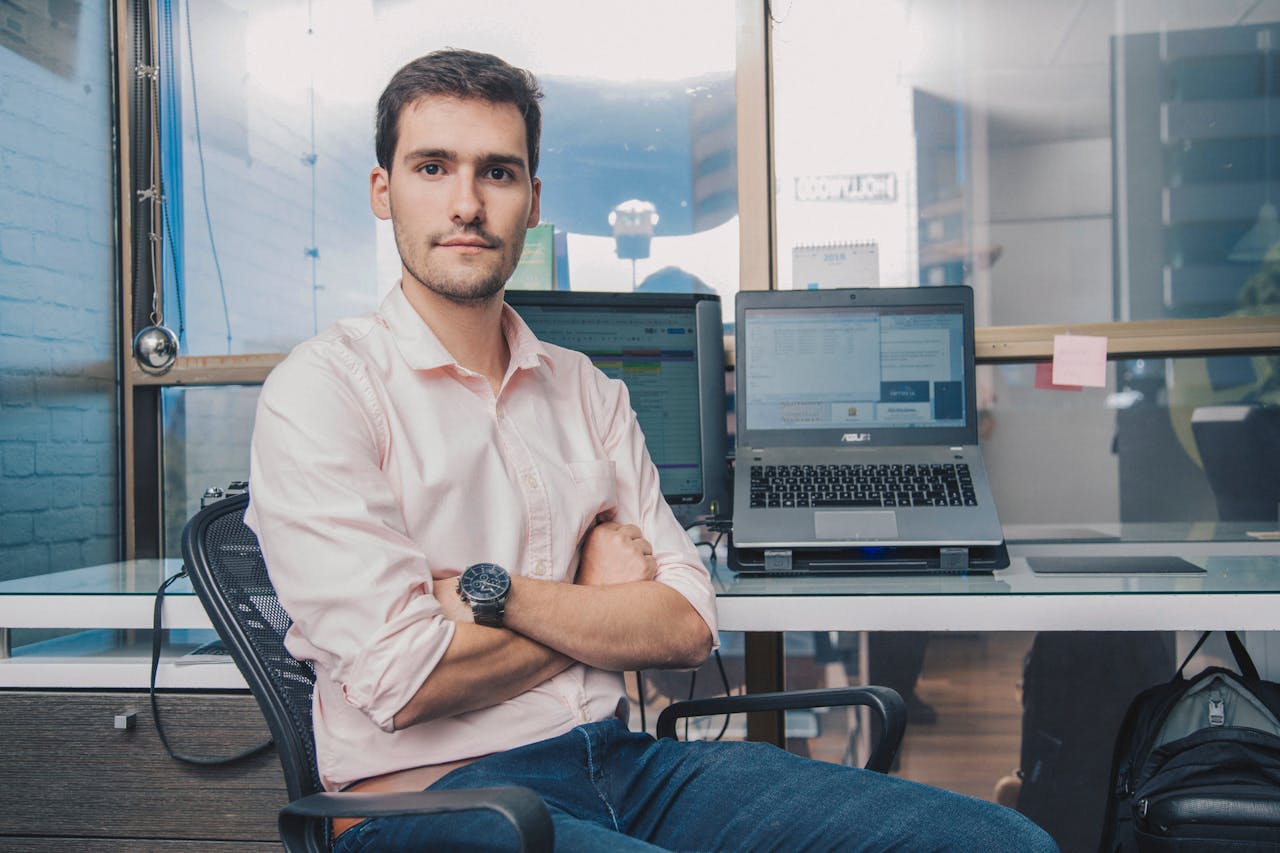 Young male professional with a confident demeanor seated at a modern office desk with a laptop.